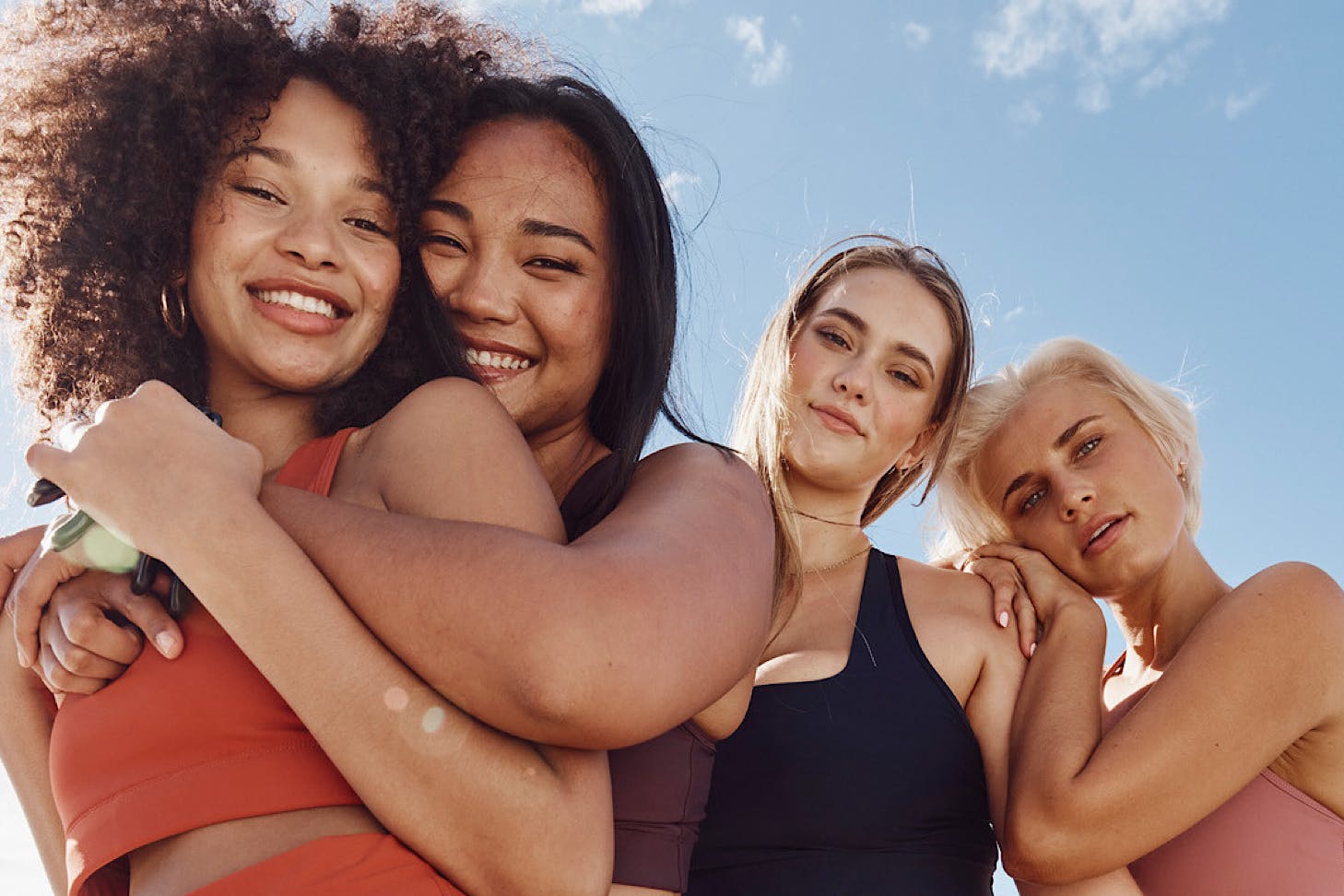 Four friends wear dk.active activewear and hug happily under a blue sky.